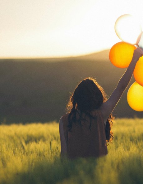 une femme heureuse de dos se tient face au soleil avec des ballons colorés dans la main