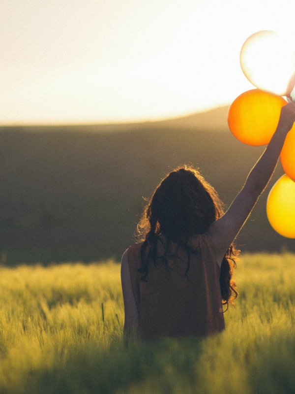 une femme heureuse de dos se tient face au soleil avec des ballons colorés dans la main